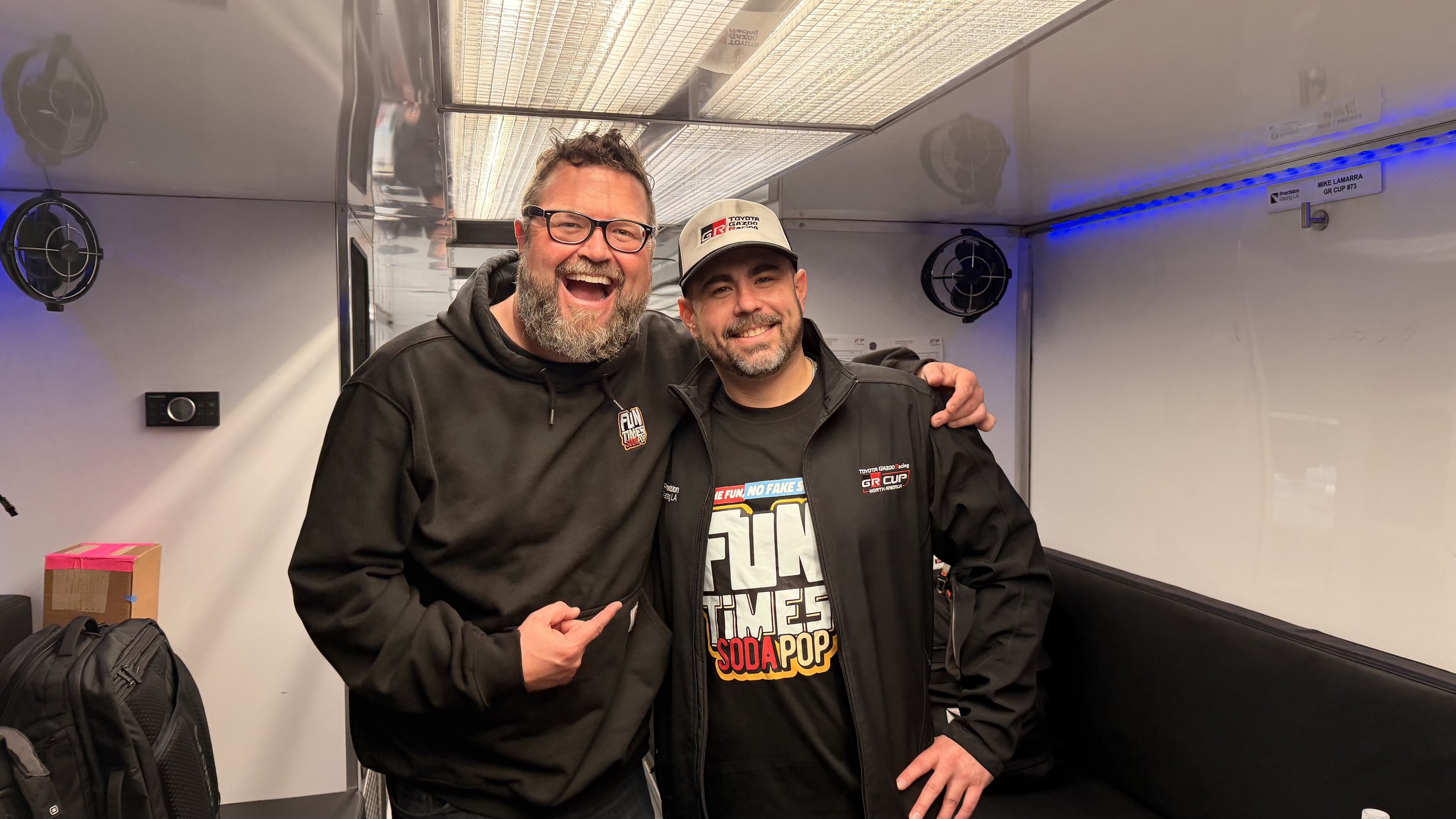 Two smiling men standing together in a room with metallic walls, one wearing glasses and a black hoodie, the other wearing a cap and a jacket with racing logos, posing with their arms around each other.