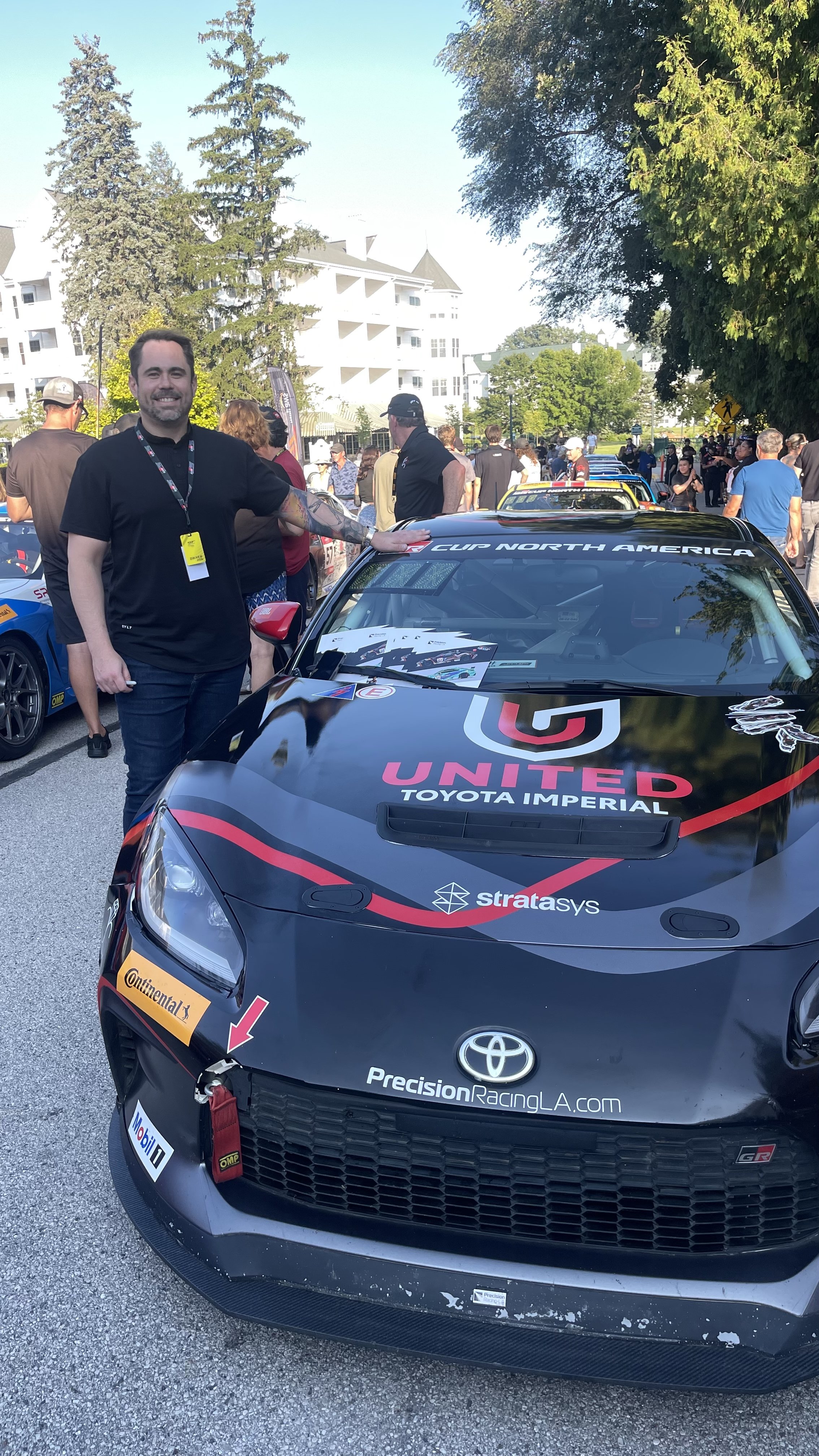 A man with tattoos and a black T-shirt standing next to a black race car with various sponsor decals at a motorsport event, with people and trees in the background.