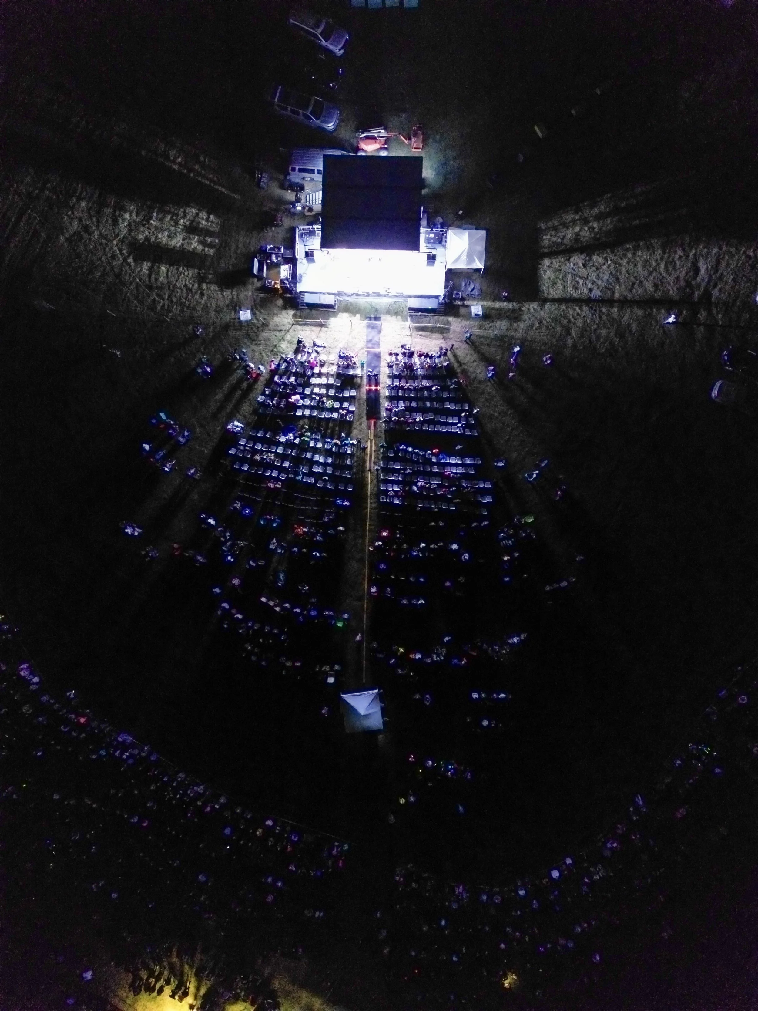 An aerial night view of a large outdoor concert or event taking place in an open field, with a brightly lit stage, rows of chairs and numerous cars parked around the area.