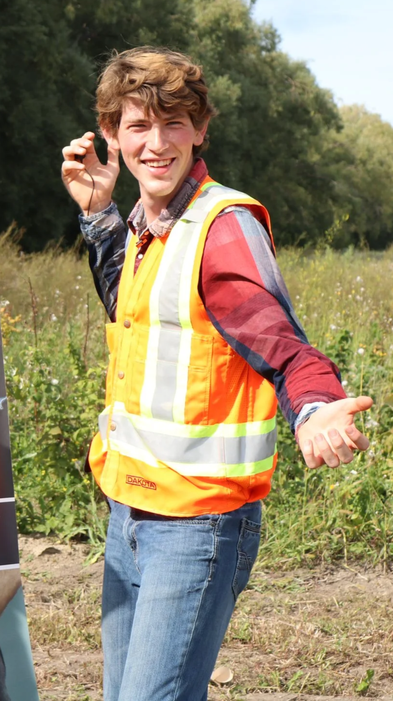 Young man smiling wearings safety vest at outdoor location with trees and grass