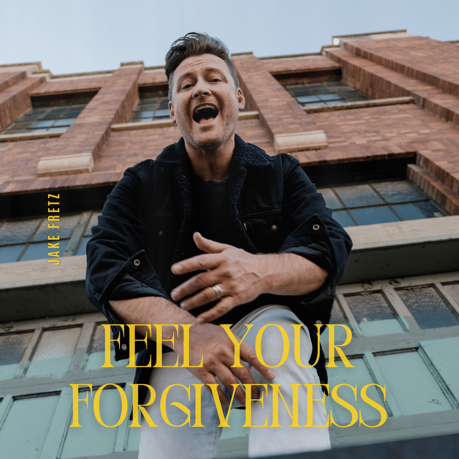A man smiling and looking down at the camera, standing below a brick building with large windows, with text reading 'Feel Your Forgiveness' and the name 'Jake Fretz'.