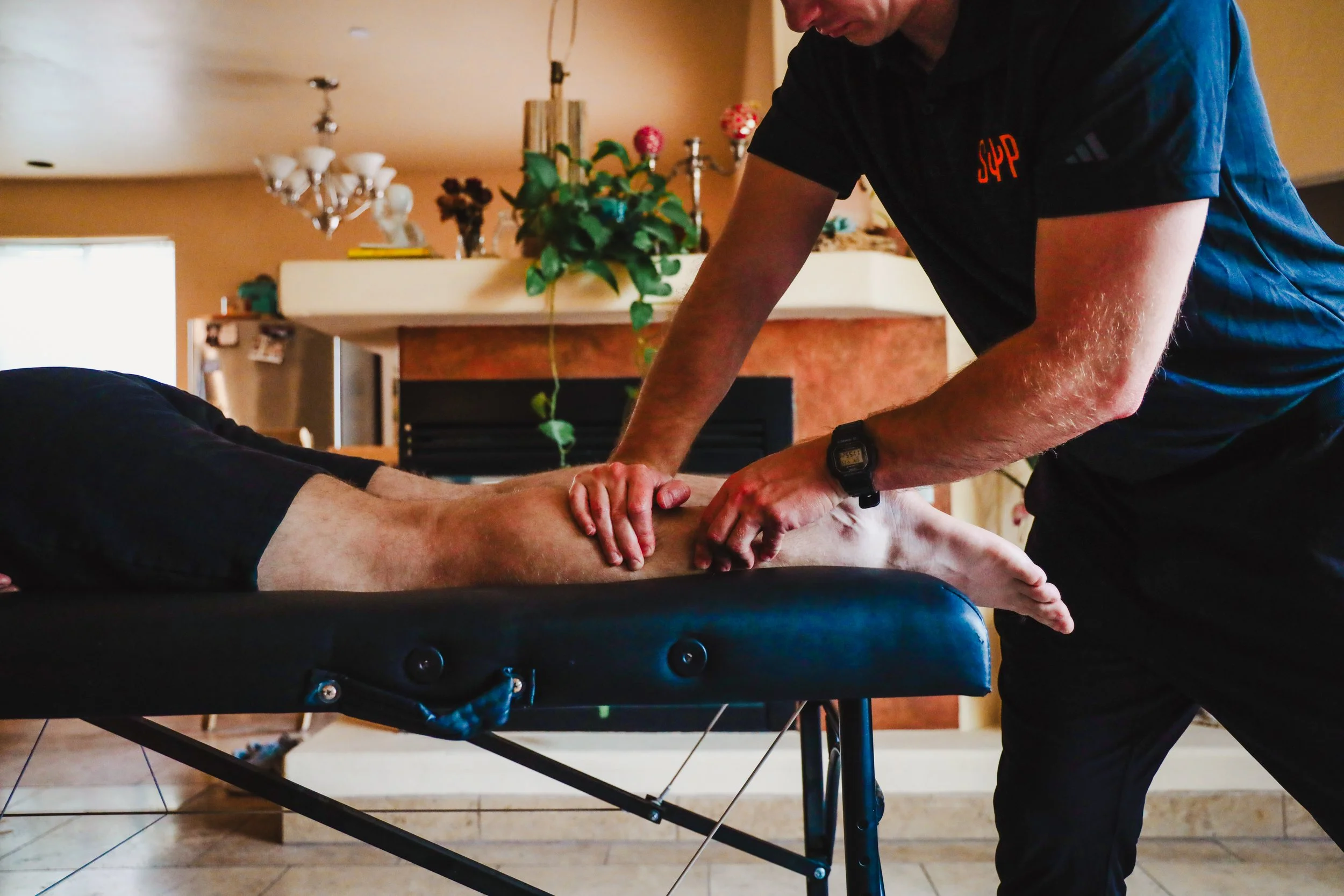 A person providing chiropractic care to a patient lying face down on a massage table, in a home setting with a fireplace and decorative plants in the background.