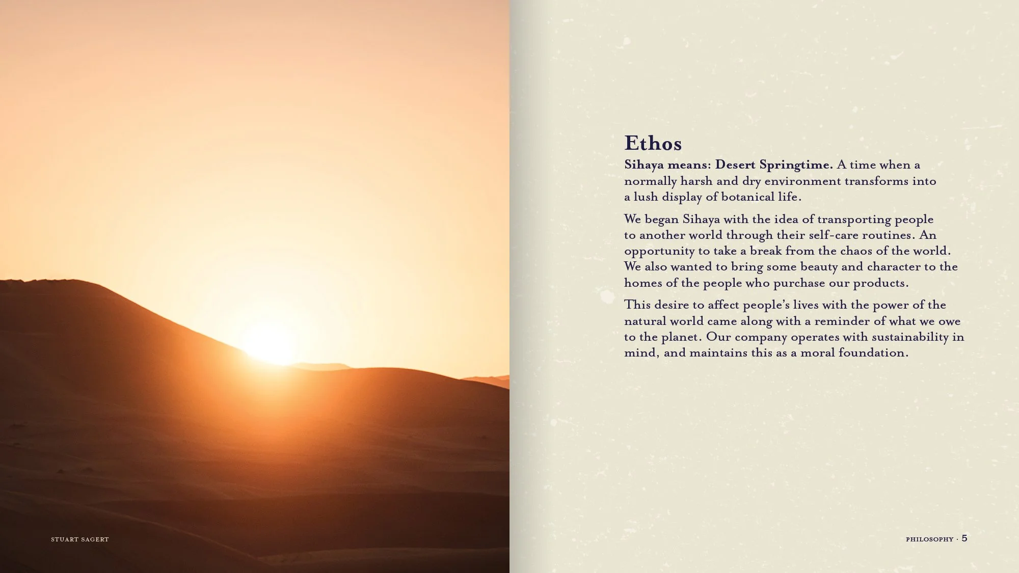 A desert landscape at sunrise with sand dunes and the sun rising behind distant mountains.