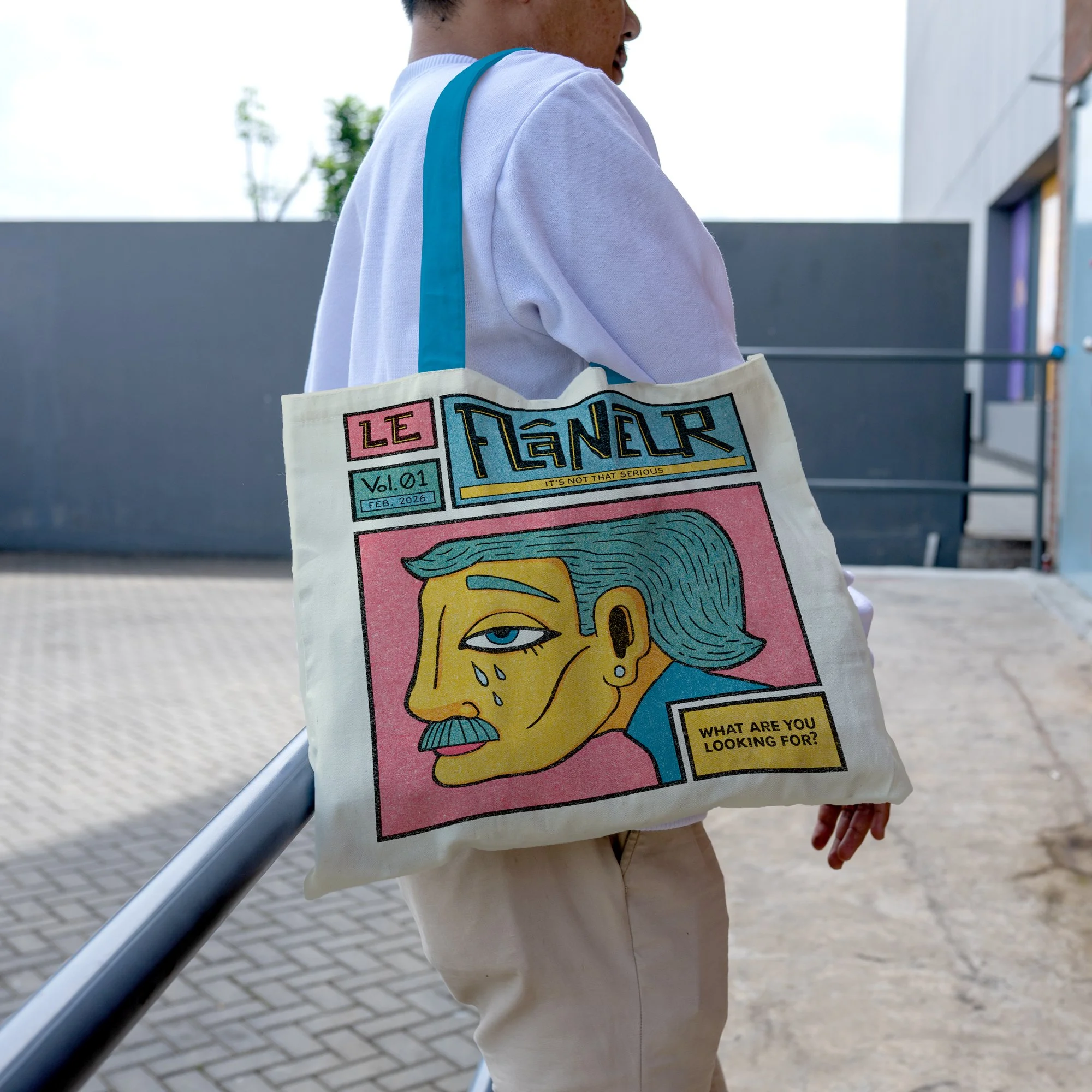 A person walking outdoors with a tote bag that features a colorful cartoon-style illustration of a man's face with blue hair, a mustache, and tears on his face. The bag has text saying "LE FLANEUR," "Vol. 01," "FEB. 2026," and "WHAT ARE YOU LOOKING FOR?" The person is wearing a white shirt and beige pants, and the tote bag has turquoise straps.