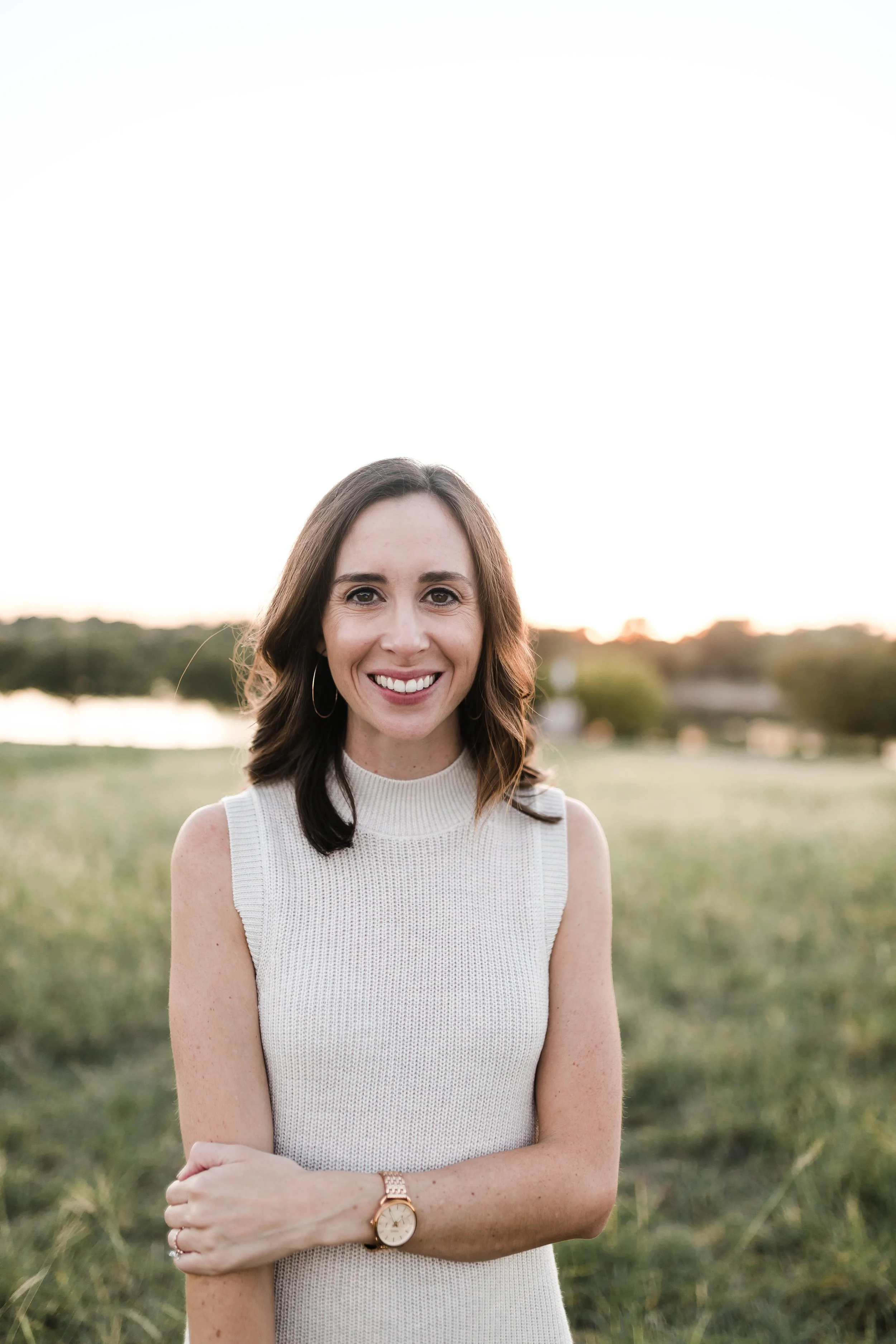 A woman standing outdoors in a field at sunset, smiling at the camera, wearing a sleeveless white top and a gold watch.