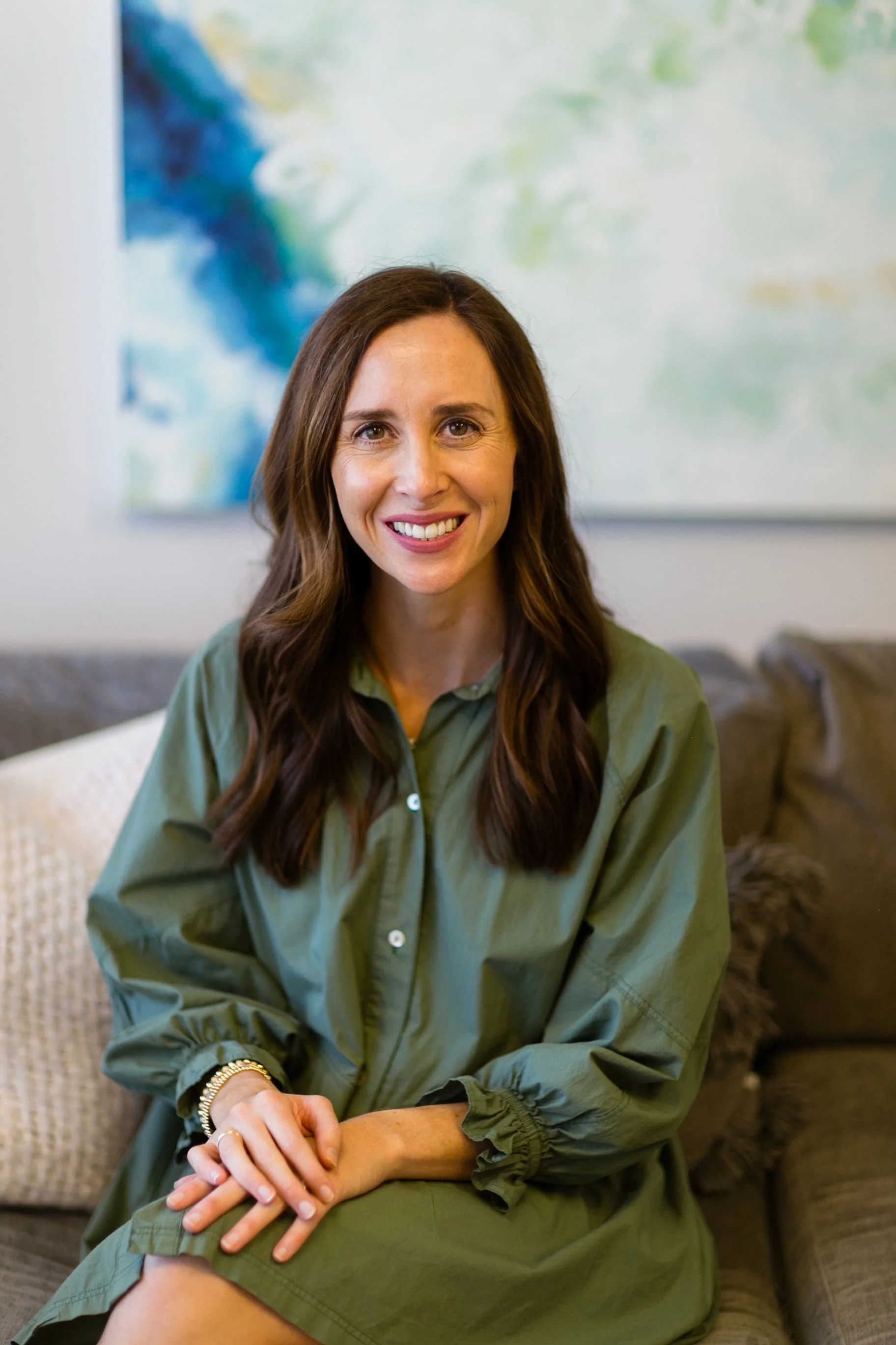 A smiling woman with long brown hair, wearing a green dress, sitting on a sofa with a beige pillow and a dark fur cushion, in a room with an abstract painting on the wall behind her.