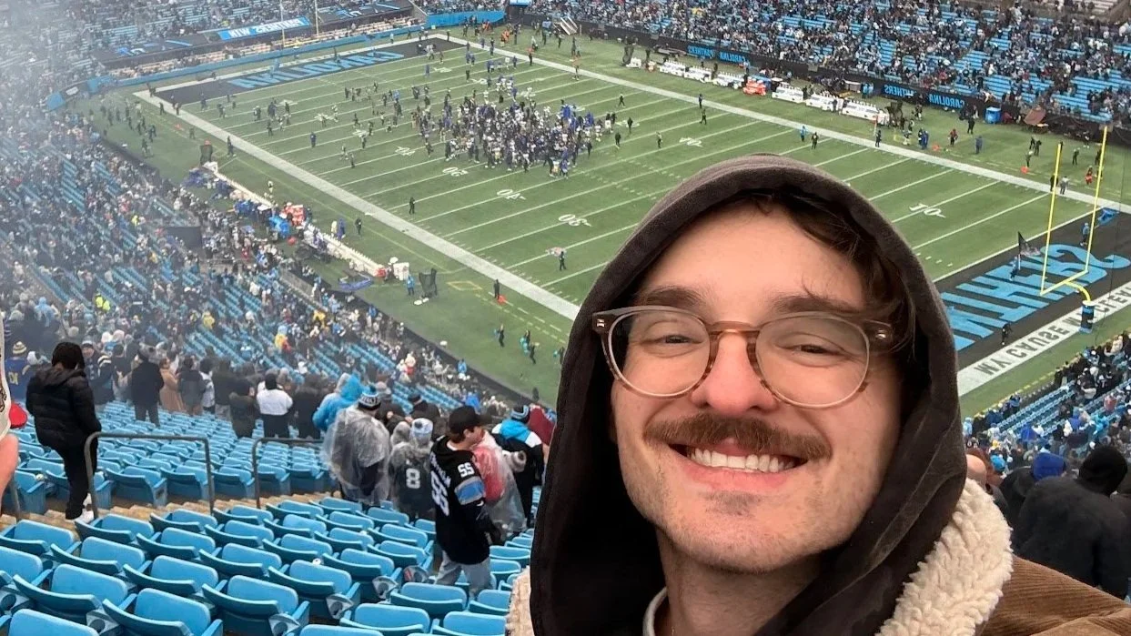 A man with glasses, a mustache, smiling in the foreground at a football stadium, with fans in the stands and players on the field in the background.