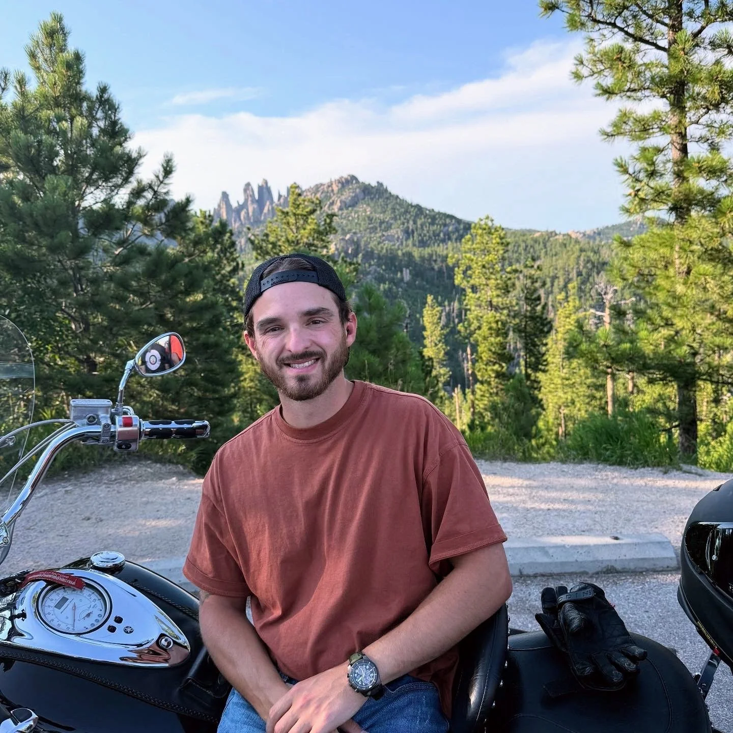 A man sitting on a motorcycle in a forested area with mountains in the background.