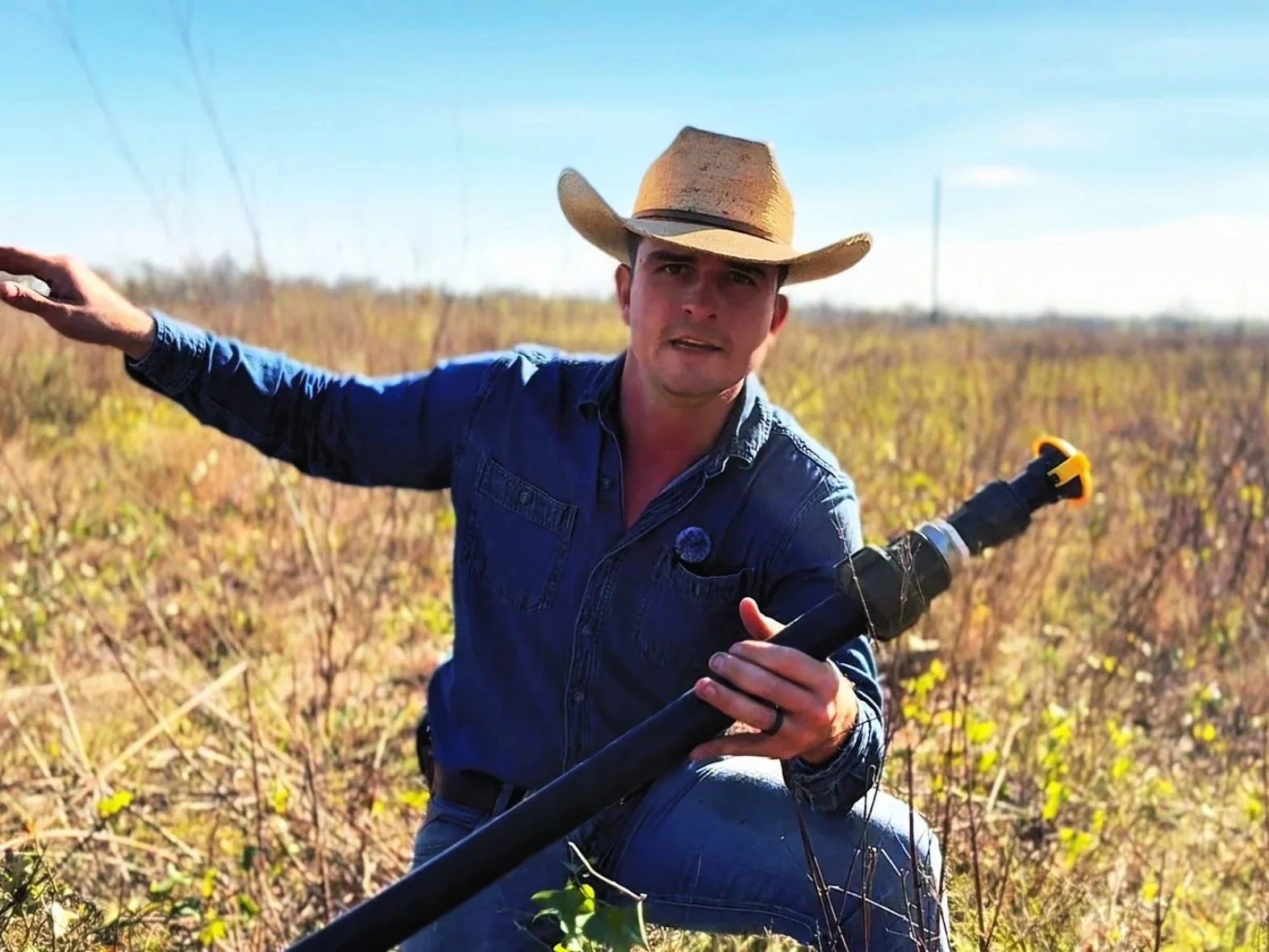 A man in a cowboy hat and a blue denim shirt kneels in a field with yellowing plants, holding a black HDPE water pipe with a yellow plasson quick connect valve, under a clear blue sky.