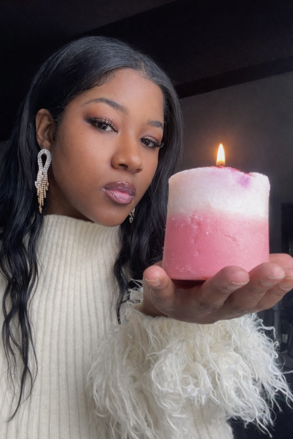A woman with dark hair and earrings holds a pink candle with a lit flame, wearing a cream-colored sweater with feathered sleeves.
