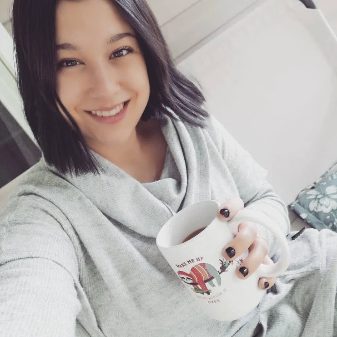 A woman with shoulder-length dark hair smiling and holding a white mug with a design, wearing a light gray hoodie.