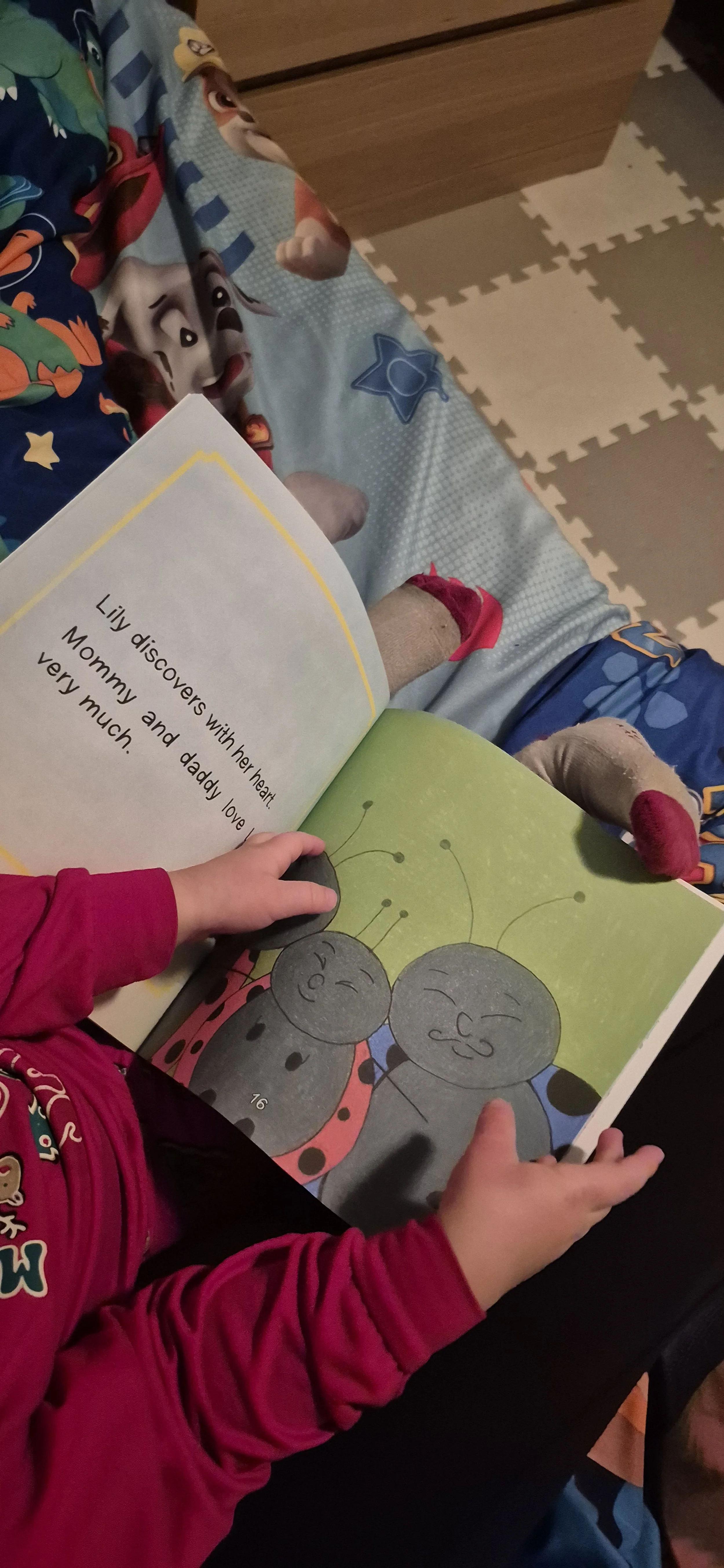 Child sitting on a bed reading a children's book with cartoon ladybugs on the page, wearing a red long-sleeve shirt.