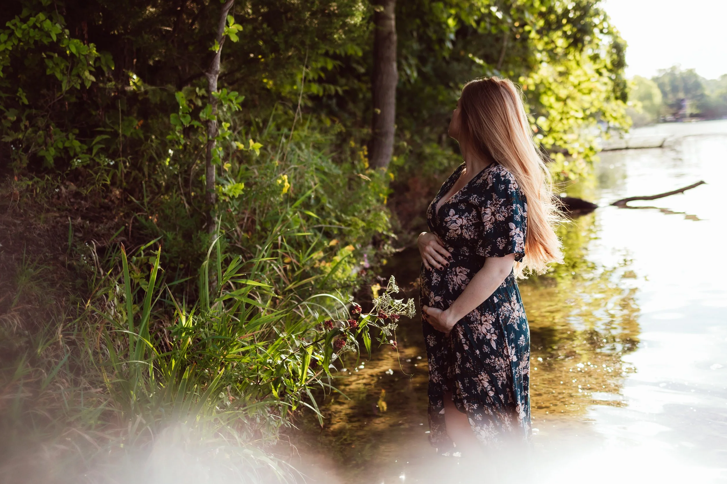 A woman with long red hair wearing a black floral dress stands in the water by a lakeshore, holding her belly, facing away from the camera, surrounded by green trees and plants.
