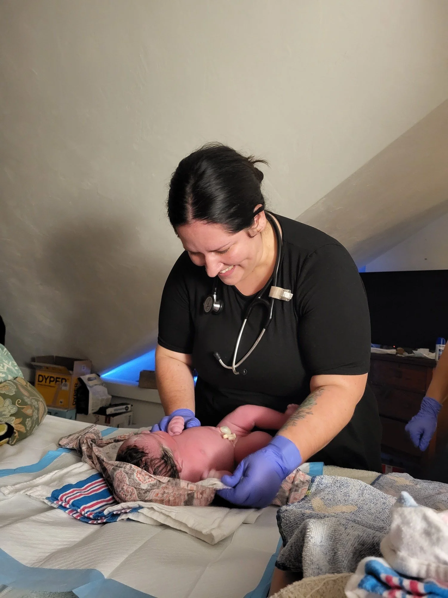 A healthcare professional, likely a nurse or doctor, caring for a newborn baby. She is smiling and wearing purple gloves, with a stethoscope around her neck.