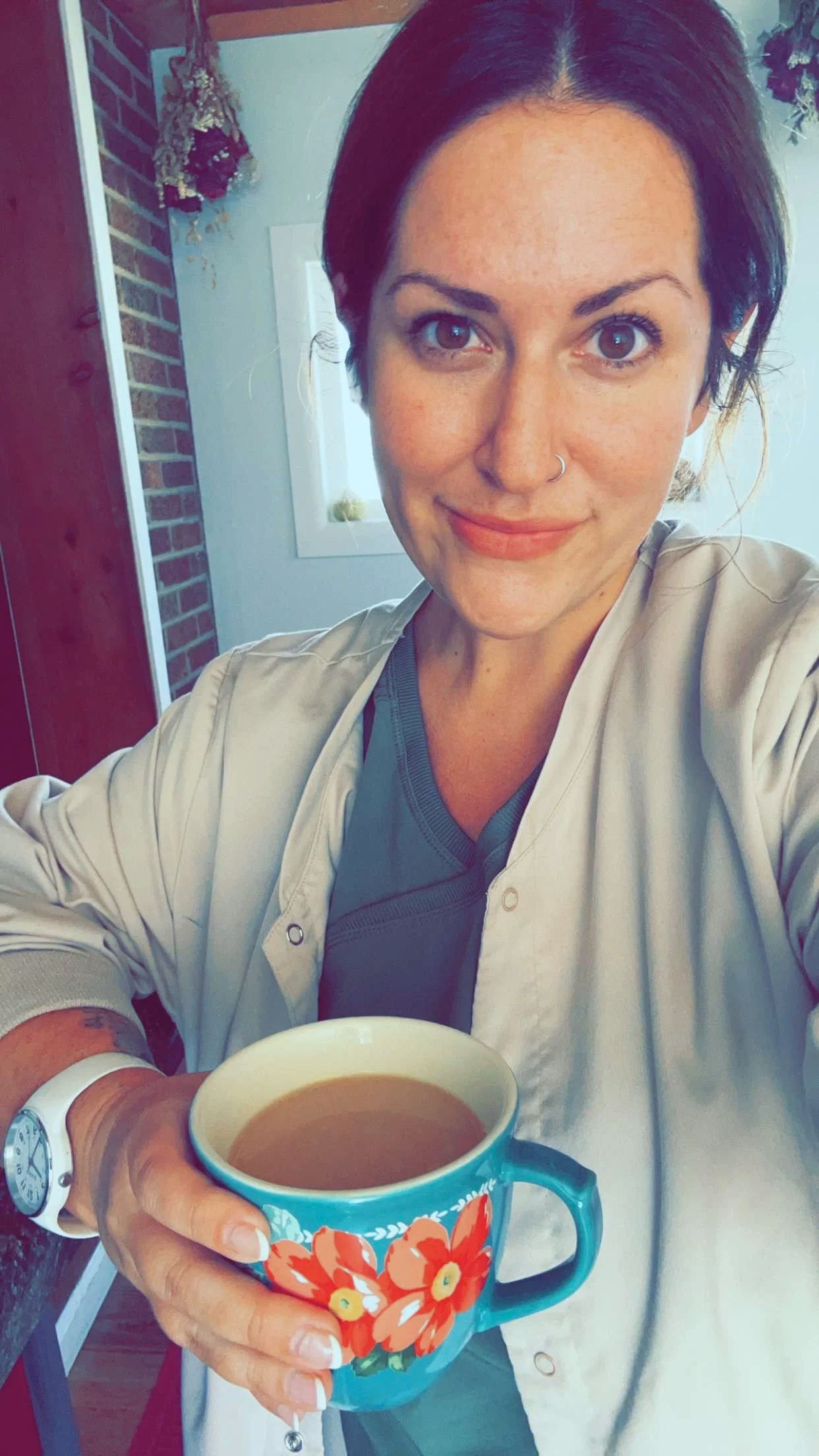 A woman with short dark hair and a nose ring holds a colorful mug filled with coffee. She is smiling and appears to be taking a selfie inside a cozy home with a brick wall and a window in the background.  She is a midwife.