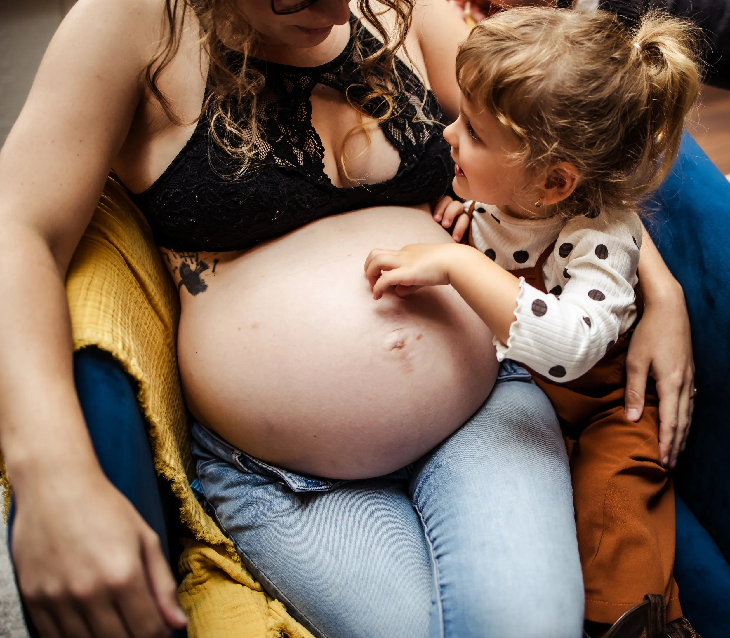 A pregnant woman sitting on a couch with a young girl touching her belly and looking at her.