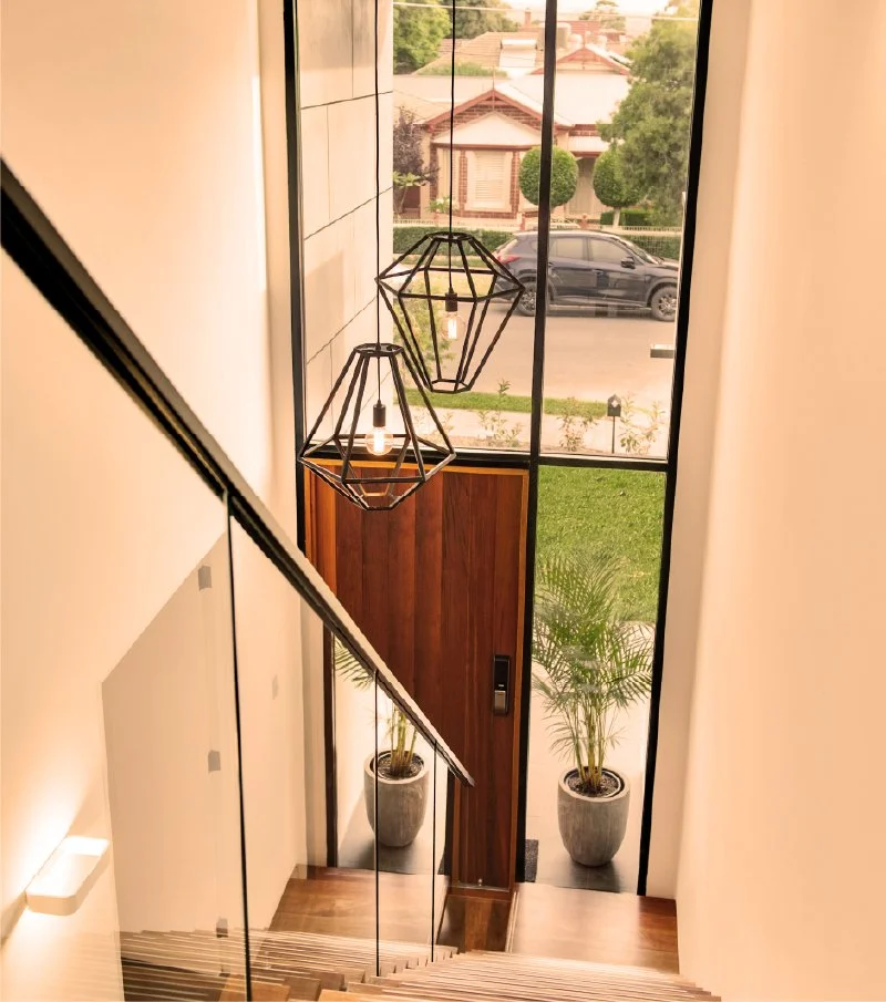 View of a residential front door with wooden paneling, flanked by two potted plants, seen from the top of a staircase inside a modern house, with pendant light fixtures hanging from the ceiling and large windows showing a street with cars and houses 
