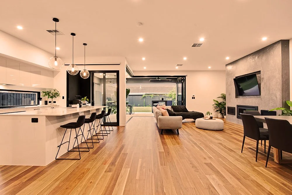 Open-concept living space with a modern kitchen on the left, a seating area with a beige and black sofa and ottomans, a wall-mounted TV, and a dining table with chairs on the right. Hardwood floors and sliding glass doors lead to an outdoor patio.