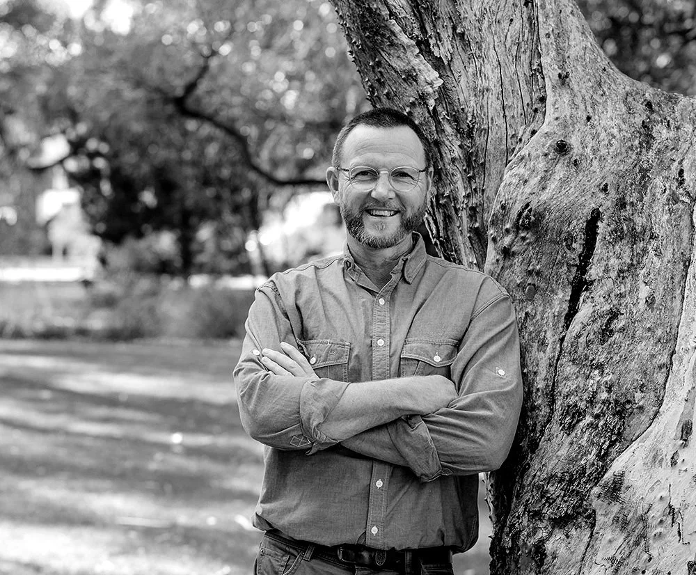 A smiling man, Ray Pollard, with glasses and a beard, wearing a button-up shirt, leaning against a tree in a park.