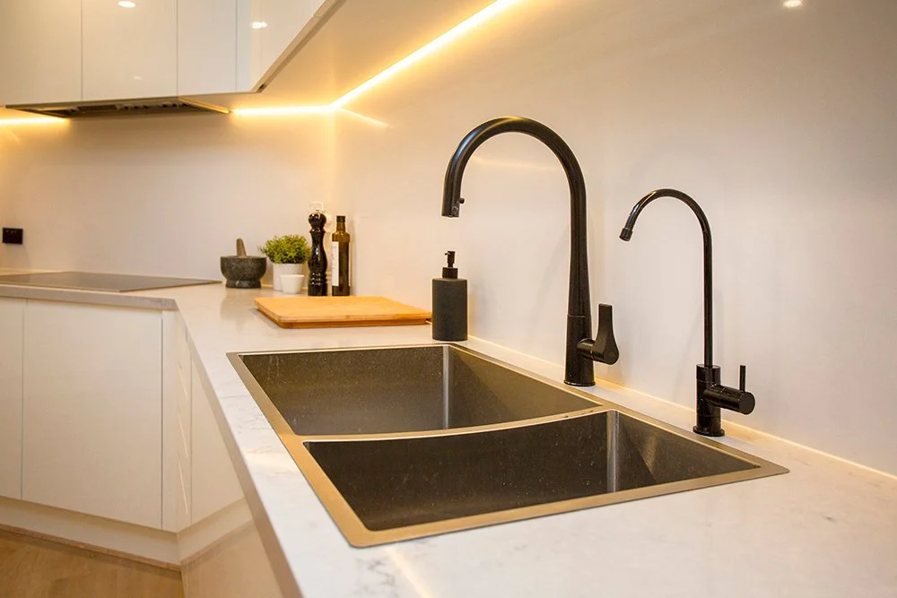 Modern kitchen countertop with dual black sinks, black faucets, soap dispenser, cutting board, potted plant, and kitchen essentials.