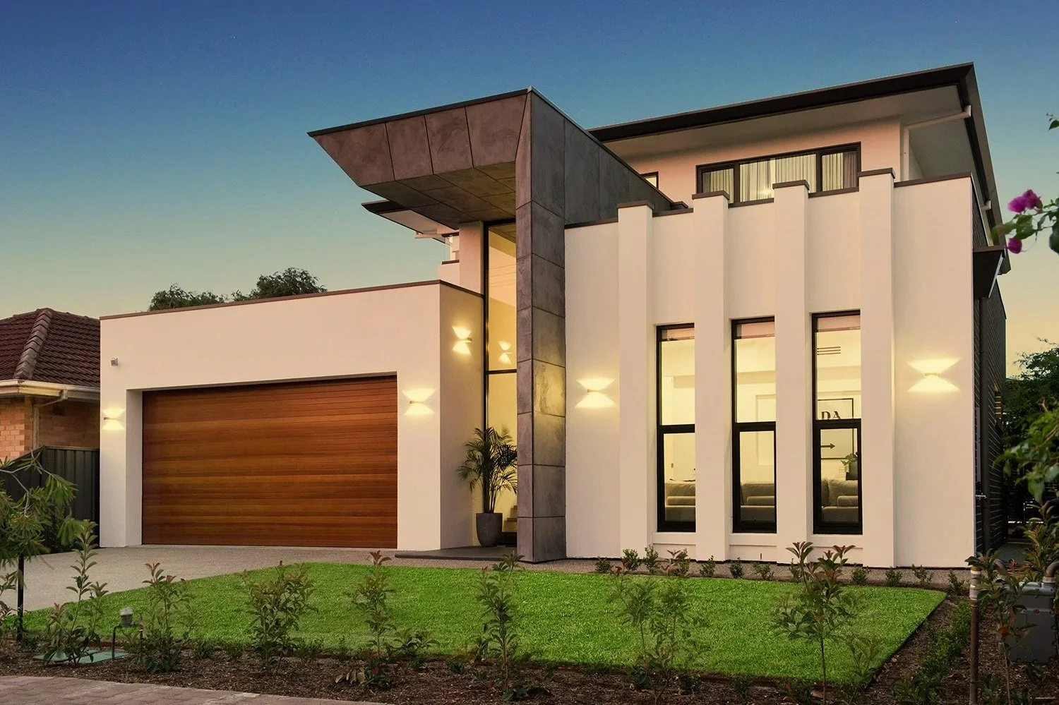  Modern two-story house with a white exterior, large windows, a wooden garage door, and a landscaped front yard with small shrubs and grass, taken during dusk with exterior lighting on.