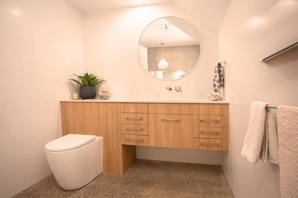 Modern bathroom with a white toilet, wooden vanity, round mirror, and decorative items on the countertop.