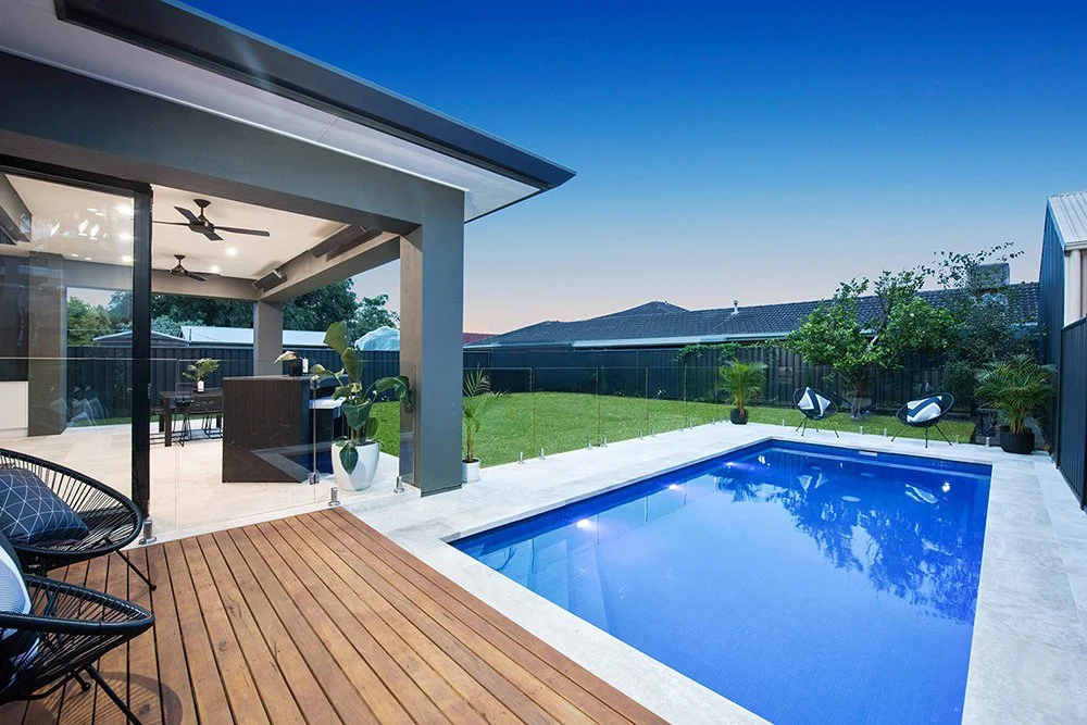 Modern backyard with a swimming pool, wooden deck, outdoor seating, and a covered patio area with ceiling fans and potted plants.