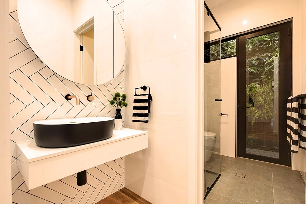 Modern bathroom with a black vessel sink on a white floating vanity, round mirror, black and brass fixtures, towel rack with black and white towel, glass shower enclosure, and a sliding glass door leading outside to greenery.