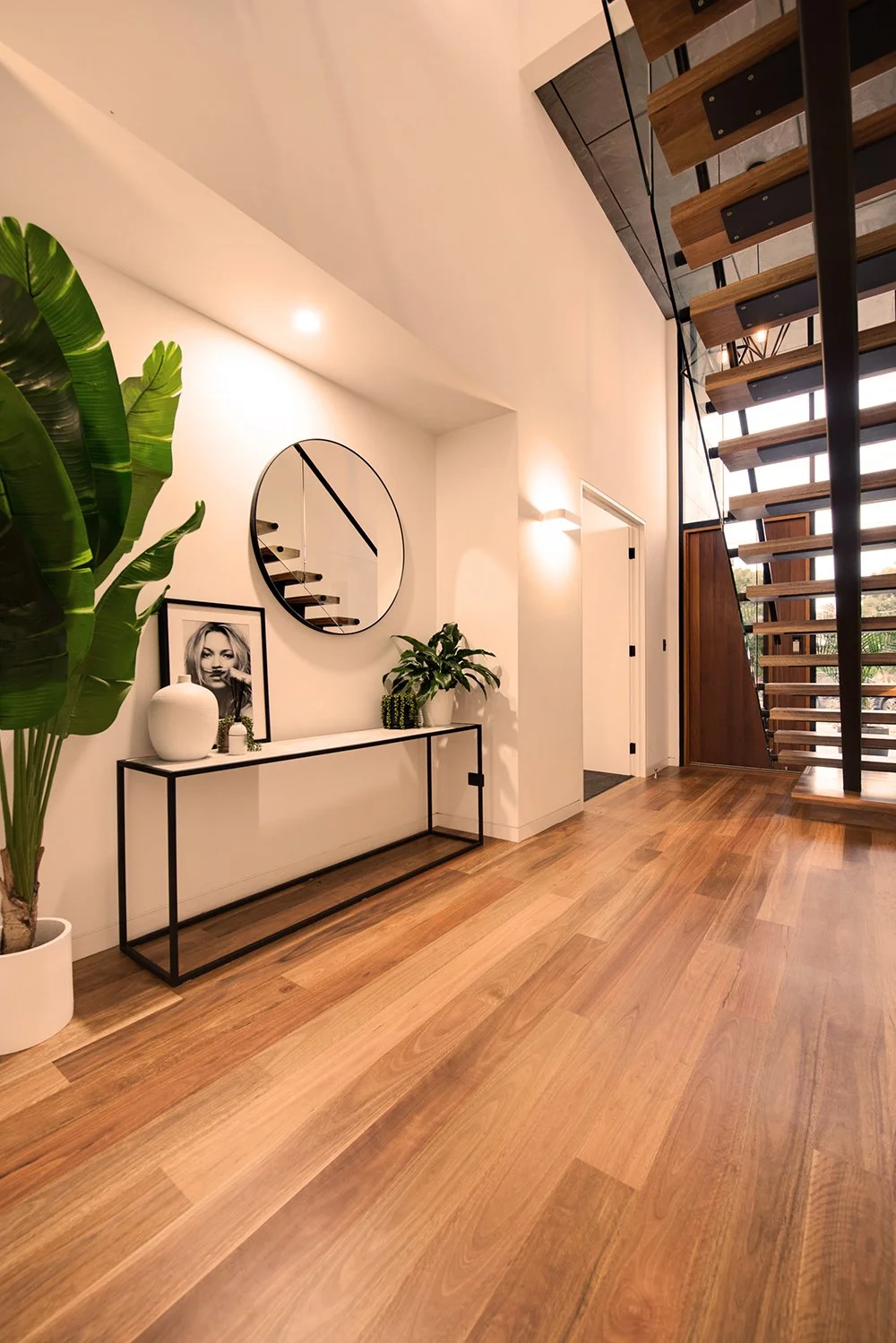 Modern interior with wooden floor, white walls, staircase with wooden steps and open risers, large window, minimalistic decor including a console table with plants and a photo, large mirror on wall, and tropical plant in a white pot.