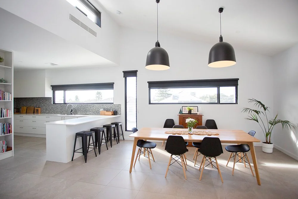 Modern open kitchen and dining area with white walls, a wooden dining table with eight black chairs, black pendant lights, a white kitchen with a long island, multiple windows, and a tall potted plant.