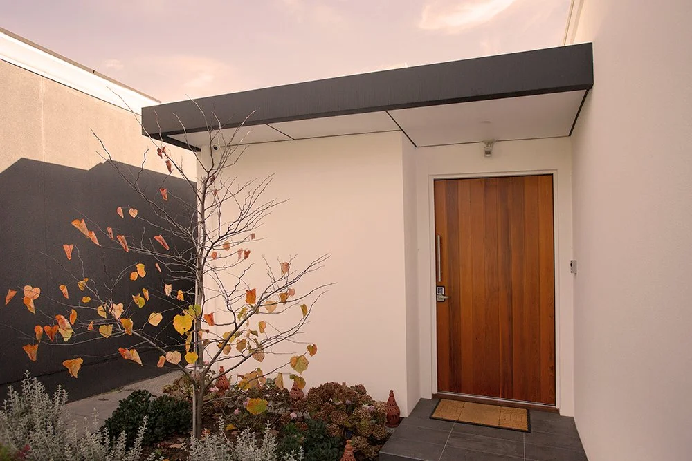 Modern house entrance with a wooden door, small garden with a leafless tree and plants, minimalist design with white and dark walls.