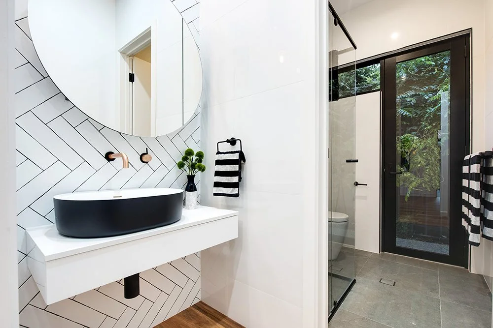 Modern bathroom with a black vessel sink on a white floating vanity, round mirror, black and brass fixtures, towel rack with black and white towel, glass shower enclosure, and a sliding glass door leading outside to greenery.