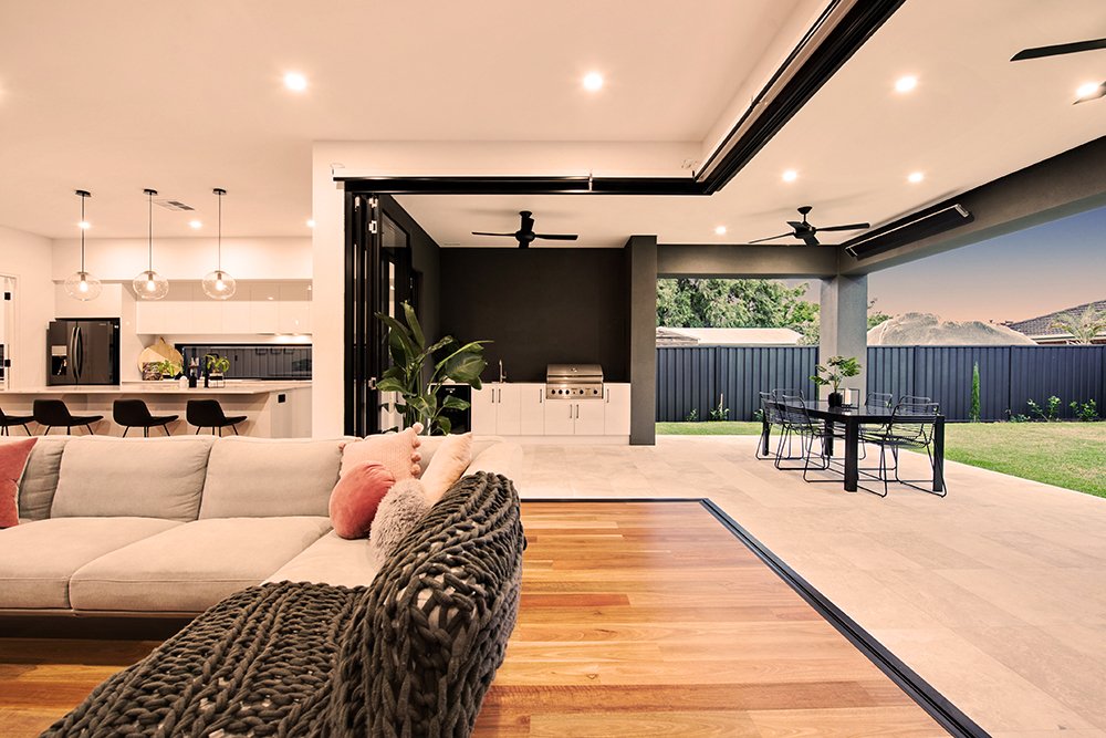 Modern open-concept living space with a white sectional sofa with pink and gray pillows, hardwood flooring, and a view of a backyard with a patio dining area and green lawn, all illuminated by ceiling lights.