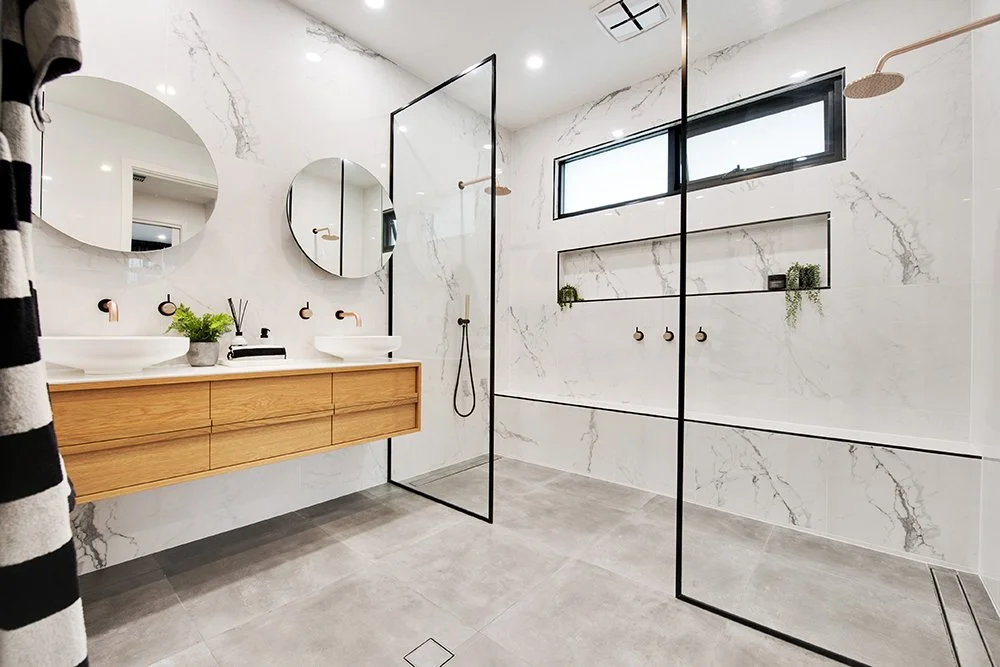 Modern bathroom with double sinks, wooden vanity, circular mirrors, gray floor tiles, marble walls, glass shower enclosure, black trim, and windows with built-in shelves.