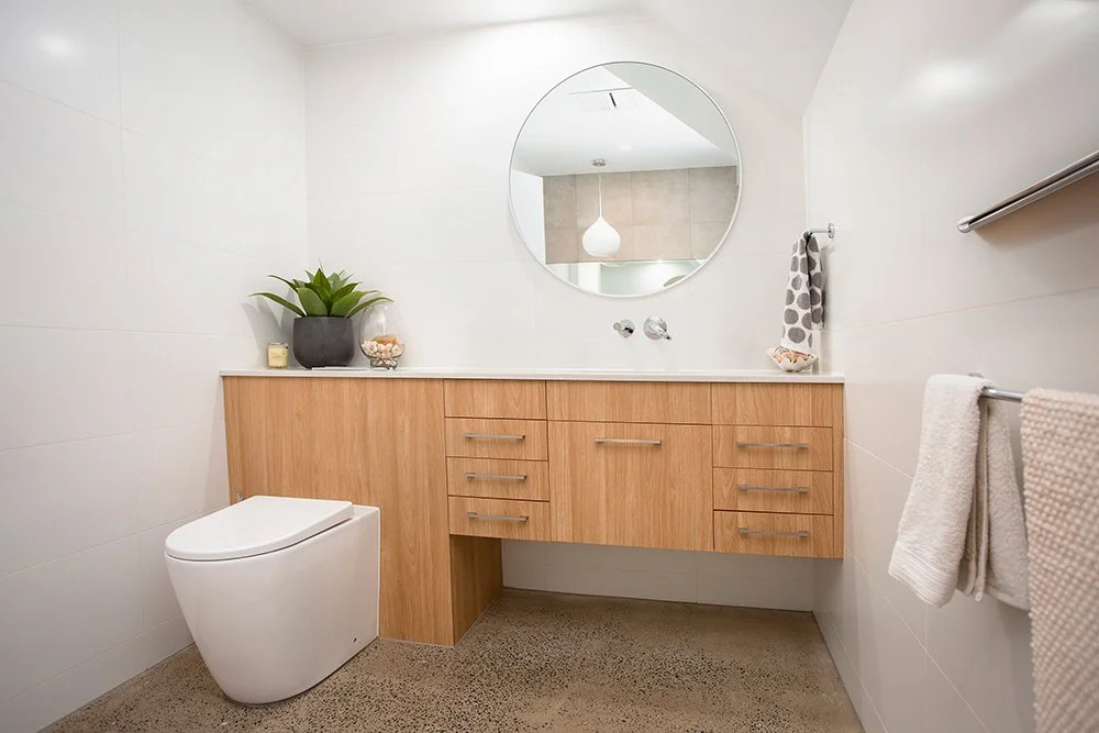 Modern bathroom with a white toilet, wooden vanity, round mirror, and decorative items on the countertop.