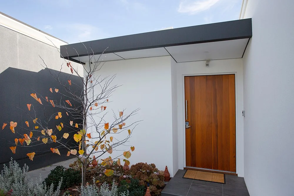 Modern house entrance with a wooden door, small garden with a leafless tree and plants, minimalist design with white and dark walls.