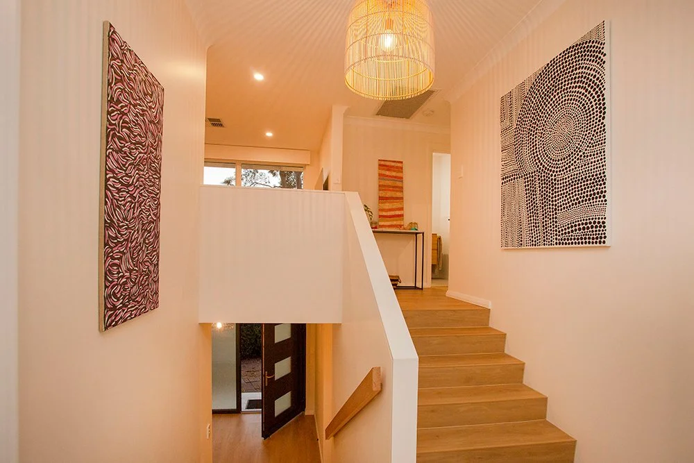 Interior of a modern home with white walls, wooden stairs, colorful abstract paintings, a hanging pendant light, and a view into another room.