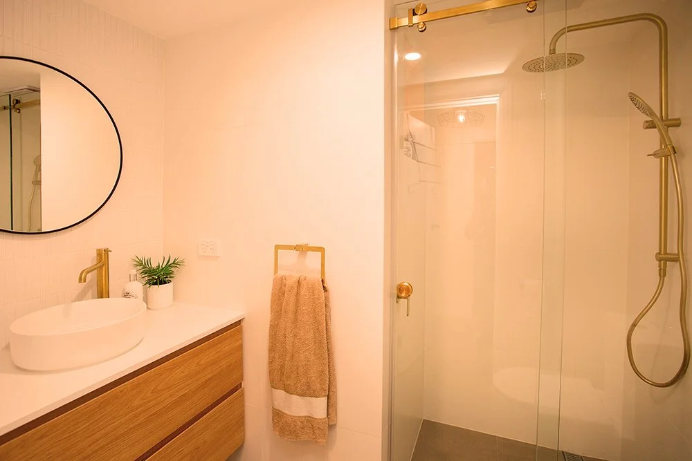 Modern bathroom with a white sink, a round mirror, a gold faucet, a towel rack with a beige towel, a glass shower with gold fixtures, and a potted plant on the countertop.