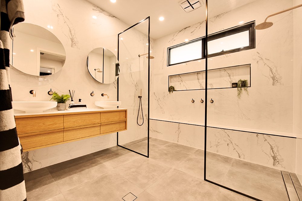 Modern bathroom with double sinks, wooden vanity, circular mirrors, gray floor tiles, marble walls, glass shower enclosure, black trim, and windows with built-in shelves.
