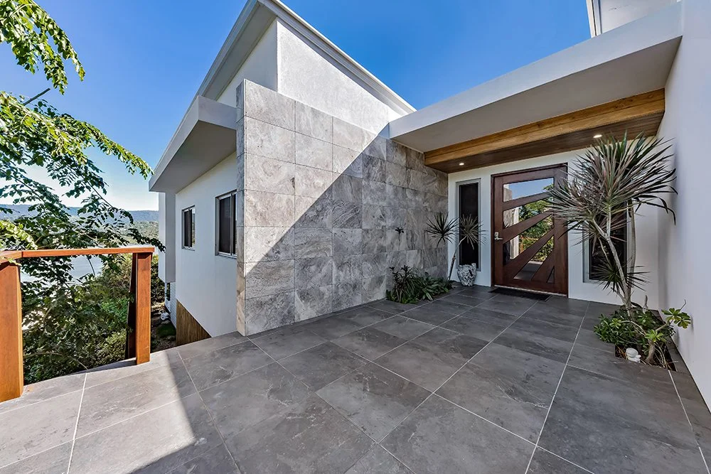 Modern house entrance with gray tiled porch, wooden front door with glass panels, potted plants, and a partial view of a wooden railing and trees outside.