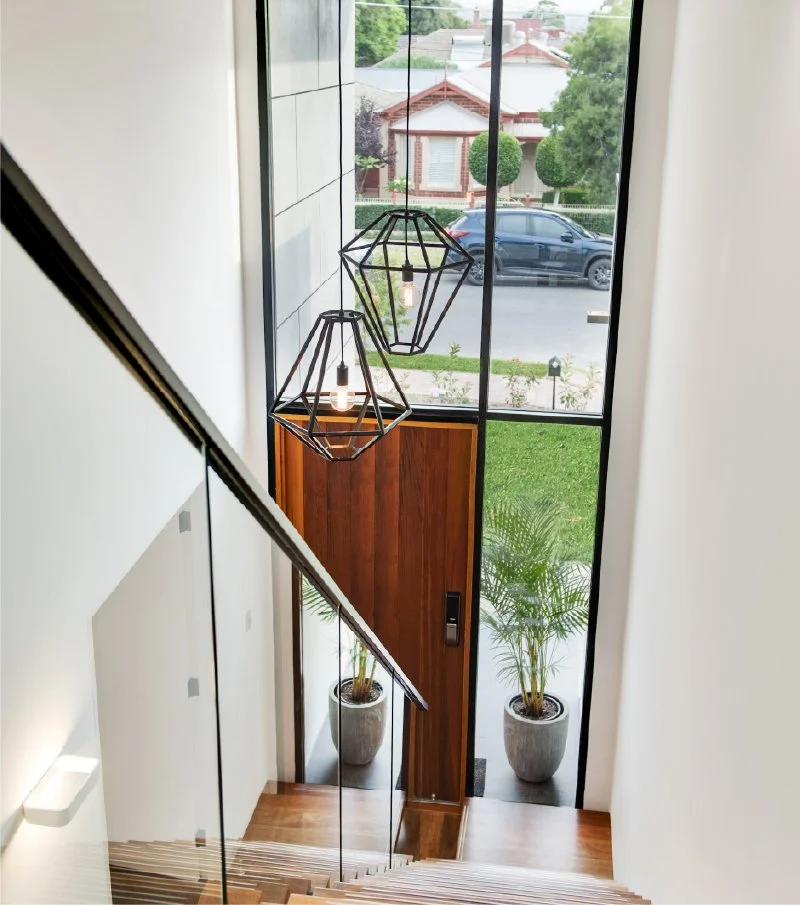 View of a residential front door with wooden paneling, flanked by two potted plants, seen from the top of a staircase inside a modern house, with pendant light fixtures hanging from the ceiling and large windows showing a street with cars and houses 