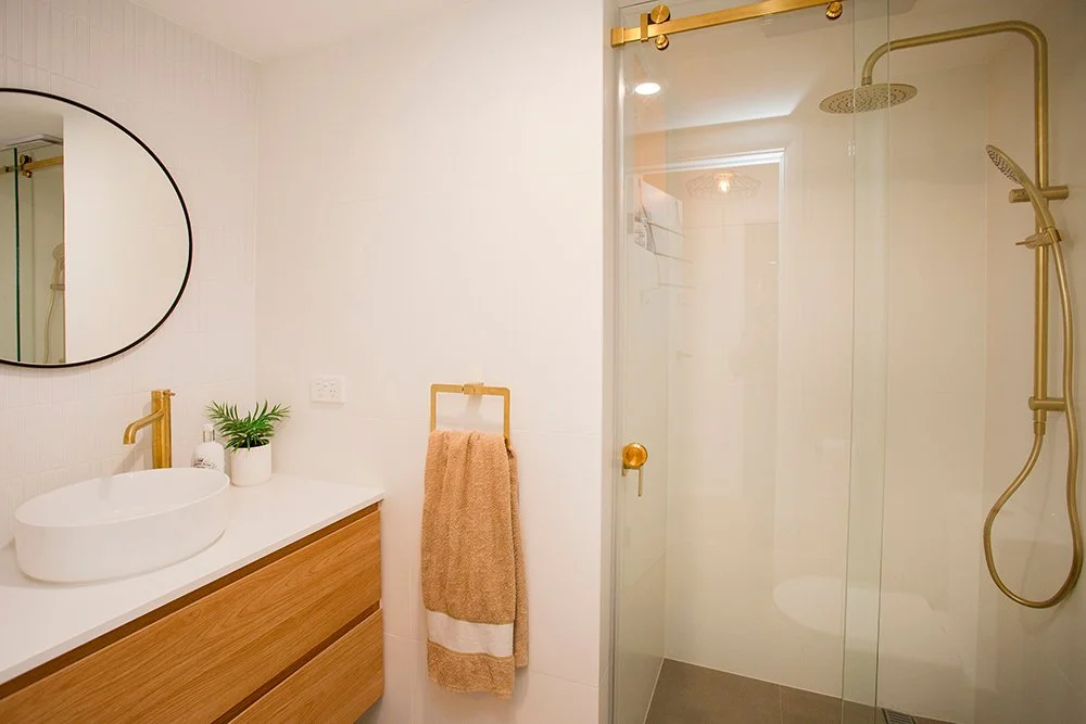 Modern bathroom with a white sink, a round mirror, a gold faucet, a towel rack with a beige towel, a glass shower with gold fixtures, and a potted plant on the countertop.