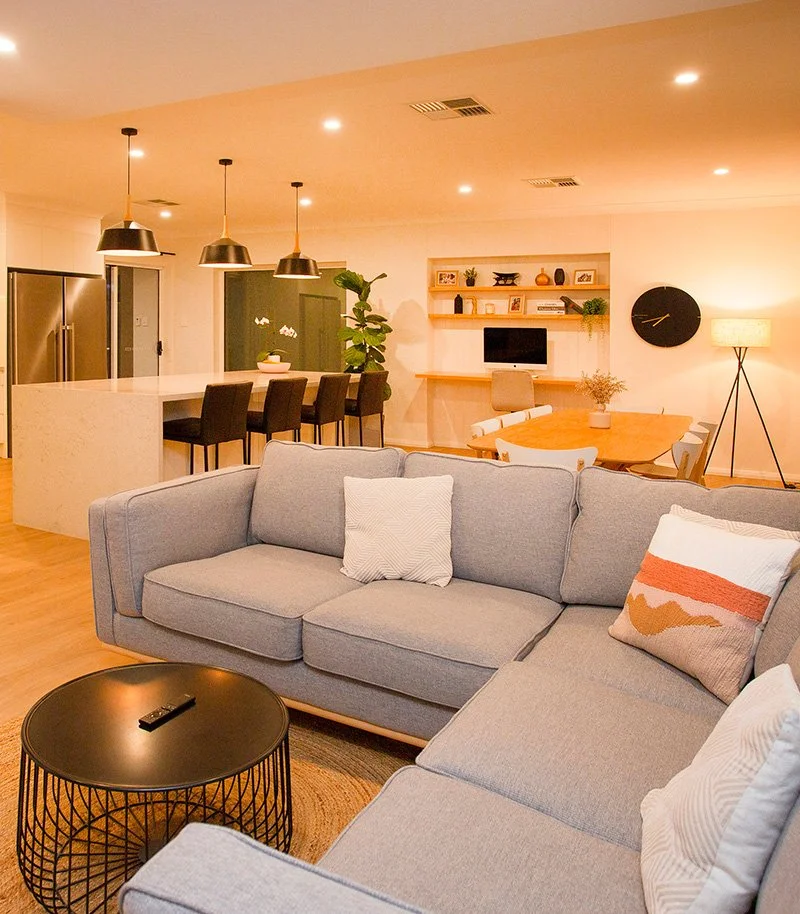 Modern open-concept living room and kitchen with a gray sectional sofa, decorative pillows, black wire coffee table, kitchen island with barstools, and a wooden dining table with chairs. Decor includes a large wall clock, shelves with decor items, an