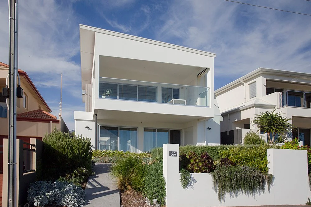 Modern white two-story house with a large balcony, glass railing, and greenery in front yard, neighboring houses visible, blue sky with clouds overhead.