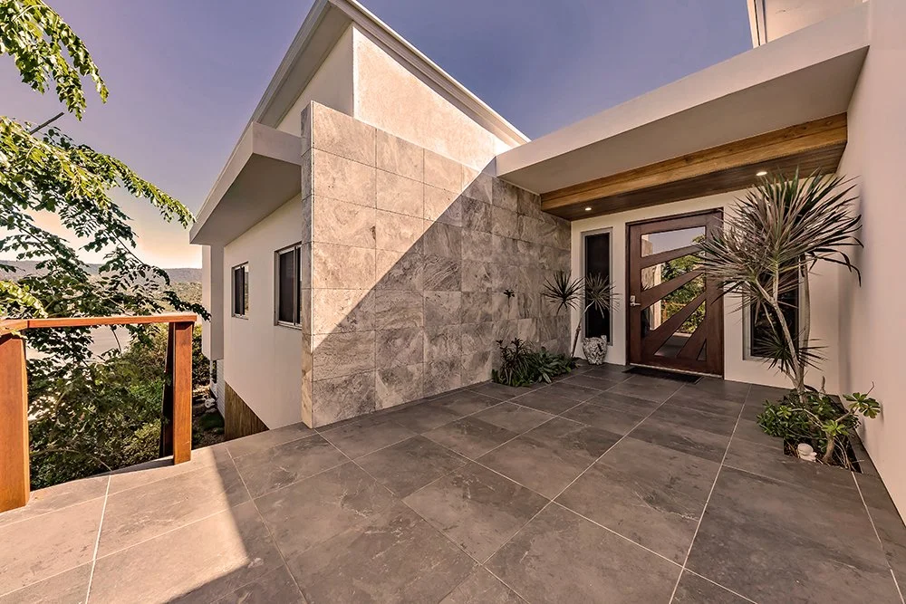 Modern house entrance with gray tiled porch, wooden front door with glass panels, potted plants, and a partial view of a wooden railing and trees outside.