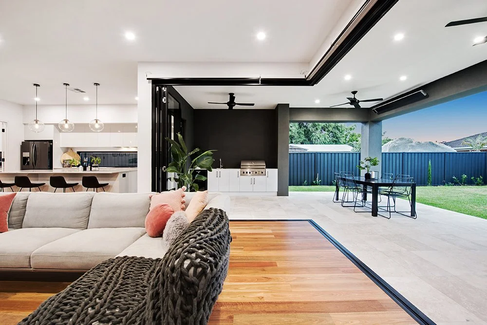 Modern open-concept living space with a white sectional sofa with pink and gray pillows, hardwood flooring, and a view of a backyard with a patio dining area and green lawn, all illuminated by ceiling lights.
