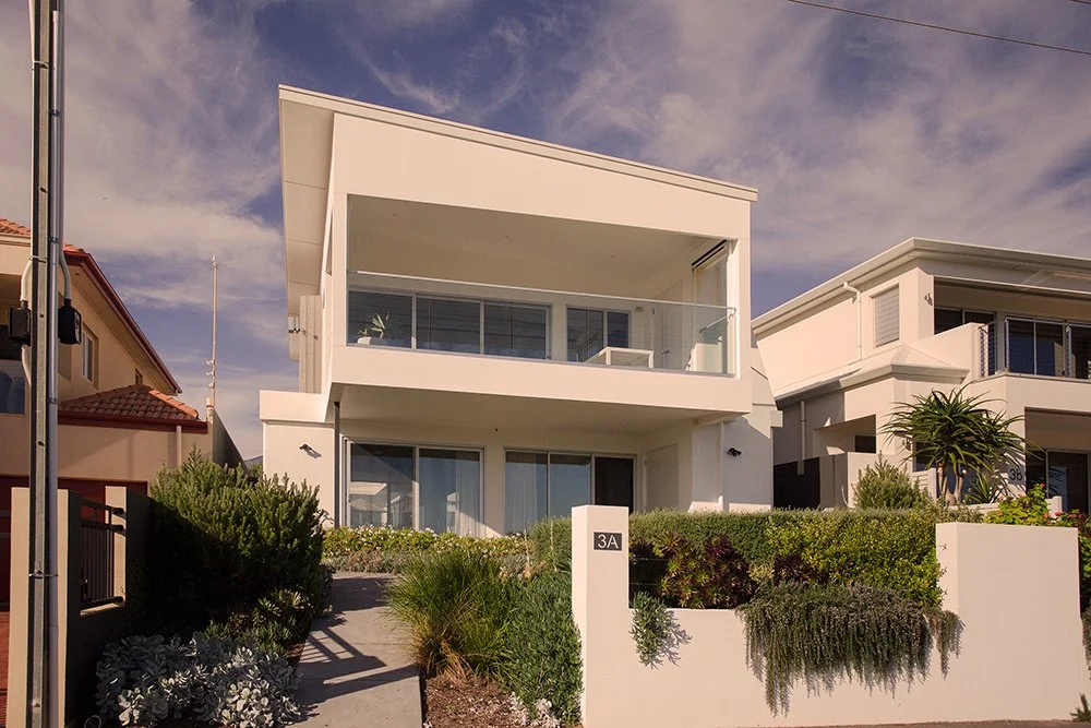 Modern white two-story house with a large balcony, glass railing, and greenery in front yard, neighboring houses visible, blue sky with clouds overhead.