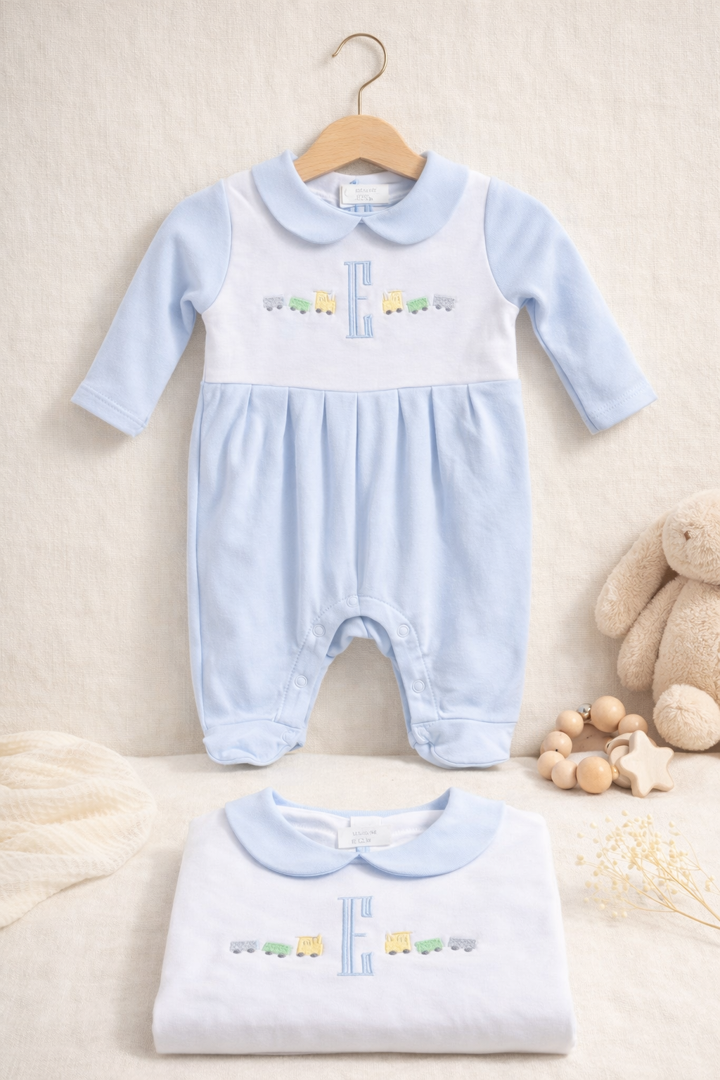 A light blue and white baby onesie hanging on a wooden hanger, and a matching folded onesie placed below on a cream surface. The clothing features a train motif and an embroidered letter "E." There are soft toys, a beaded bracelet, and decorative ele