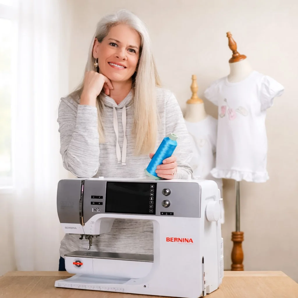 A smiling woman with long gray hair standing in a bright room, holding a spool of thread, with a sewing machine on the table in front of her and mannequins in the background wearing white clothing.