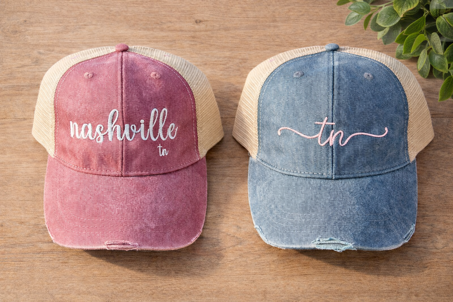 Pink and blue trucker hats with embroidered white and pink text on the front panels, placed on a wooden surface with greenery in the background.